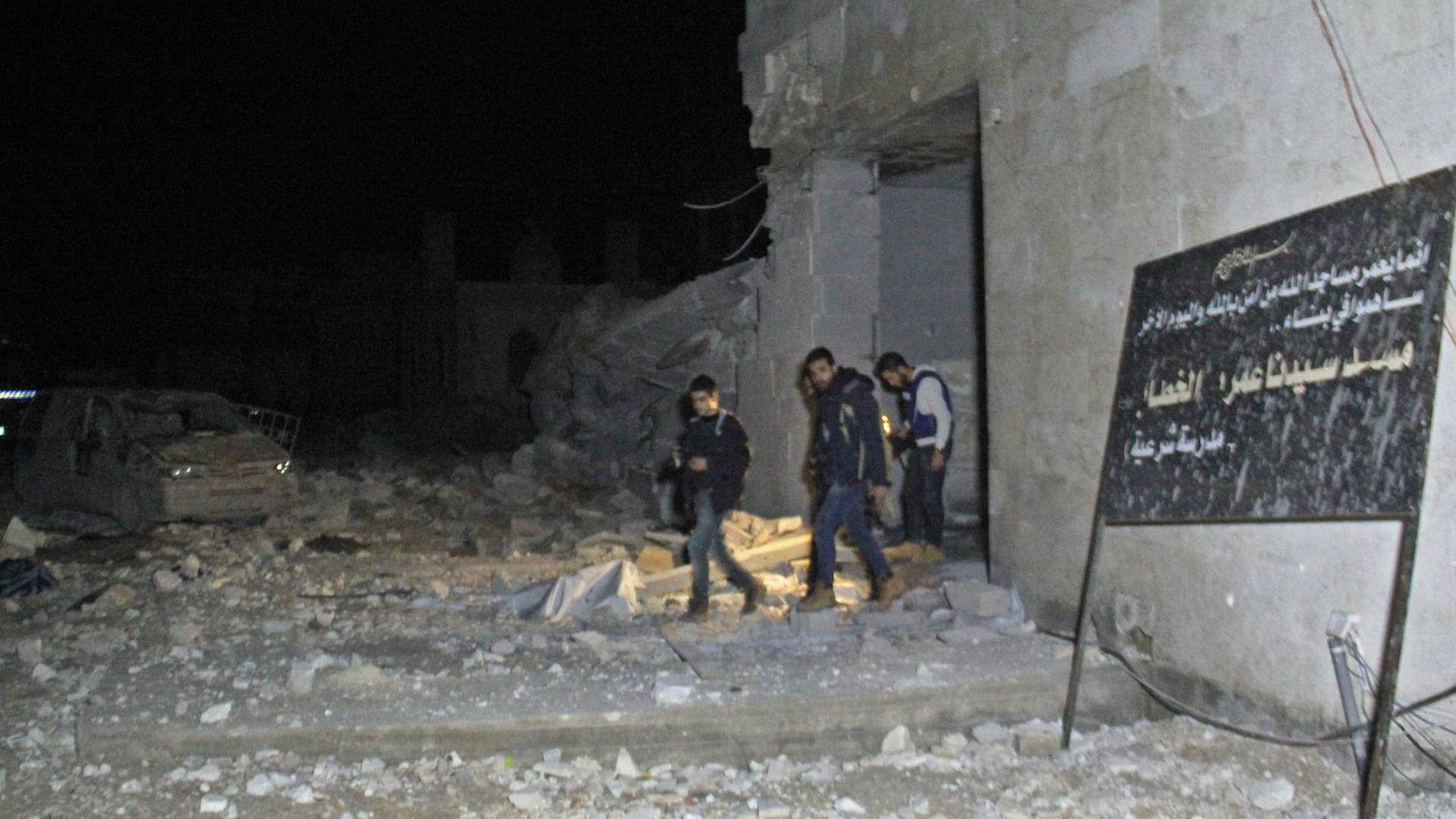 Mehrere Menschen treten aus einem halb-zerstörten Gebäude. Im Vordergrund ein Schild mit arabischer Schrift.