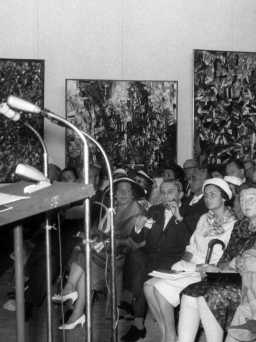 Der Kunsthistoriker Werner Haftmann steht während der Eröffnung der documenta 2 im Jahr 1959 am Rednerpult, im Publikum unter anderem sein Kollege Arnold Bode.