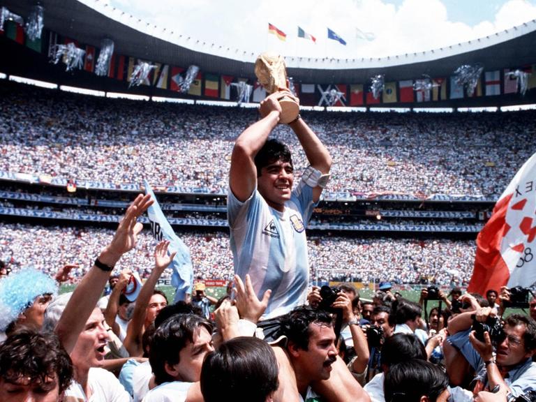 Diego Maradona wird auf den Schultern getragen und hält jubelnd den Weltmeisterpokal 1986 in der Mitte des Stadions hoch.