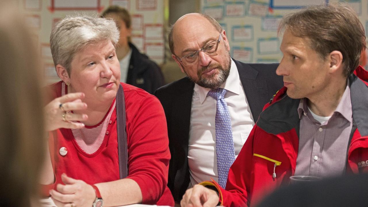 Schulz sitzt in einem Raum hinter einem Mann und einer Frau. Die Frau redet, der Mann und Schulz hören zu. Dahinter unscharf zwei Stellwände mit Organisations-Diagrammen.