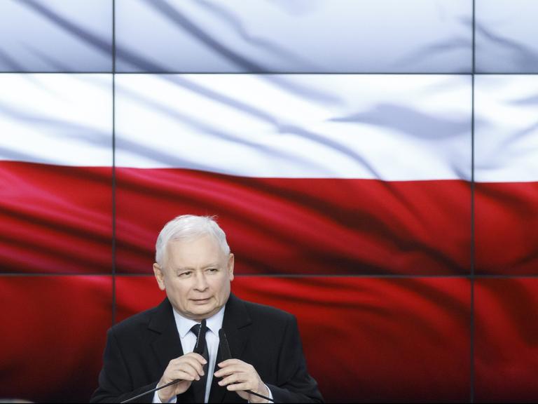 Jaroslaw Kaczynski hinter zwei Mikrofonen, im Hintergrund die polnischen Landesfarben