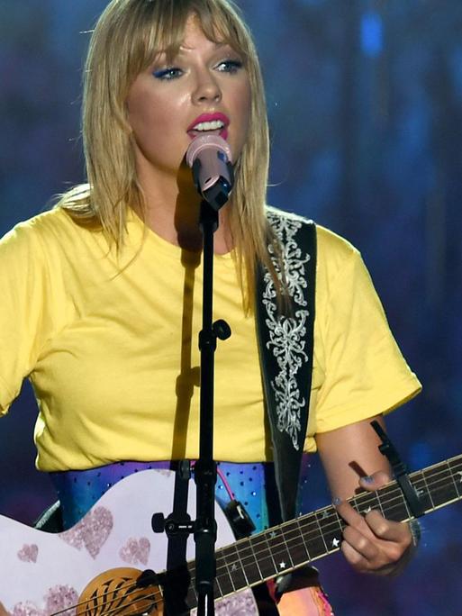 Das Bild zeigt Taylor Swift während eines Konzerts wie sie singt und dabei auf einer rosa Gitarre spielt.