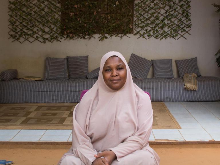 Die Predigerin Malama Sakina, in mauvefarbenes Tuch und Kopftuch gehüllt, sitzt in ihrem Haus vor einer mit Vorhängen gerahmten Nische, in der unter dem Fenster ein Sofa mit vielen kleinen Kissen steht.