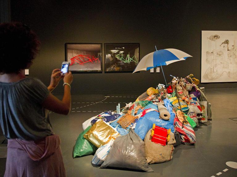 Eine Besucherin macht ein Foto von einem Ausstellungsstück von Andreas Slominski. Zu sehen sind gefüllte Müllsäcke, Tüten, Beutel, Alltagsgegenstände wie einen Teppich und Spielzeug, die auf einem Haufen liegen. Ganz oben steckt ein Regeschirm. Das Ganze ist der Teil der Ausstellung, die 2013 in Barcelona gezeigt wurde.