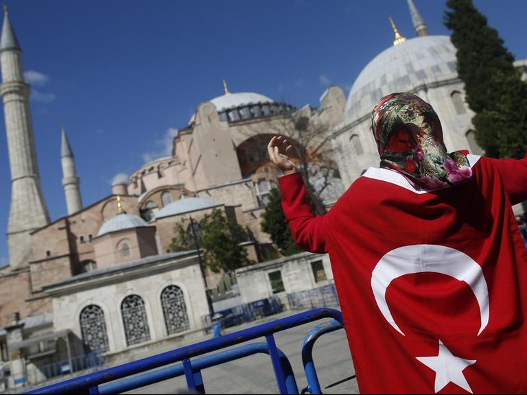 Eine Frau ist in eine türkische Flagge gewickelt und hebt die Hände gen Himmel. Vor ihr die Hagia Sophia.