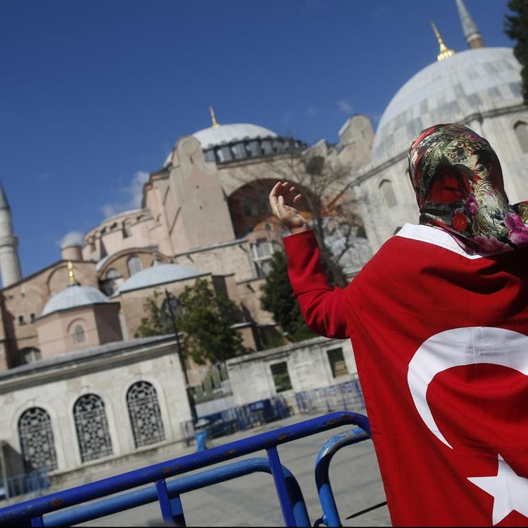 Eine Frau ist in eine türkische Flagge gewickelt und hebt die Hände gen Himmel. Vor ihr die Hagia Sophia.