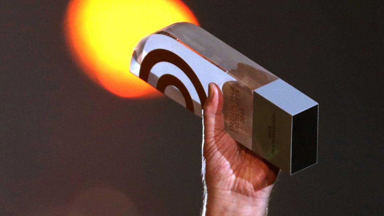 Eine Hand reckt die Trophäe für Gewinner des Deutschen Radiopreises nach oben, im Hintergrund ein verschwommenes Scheinwerferlicht