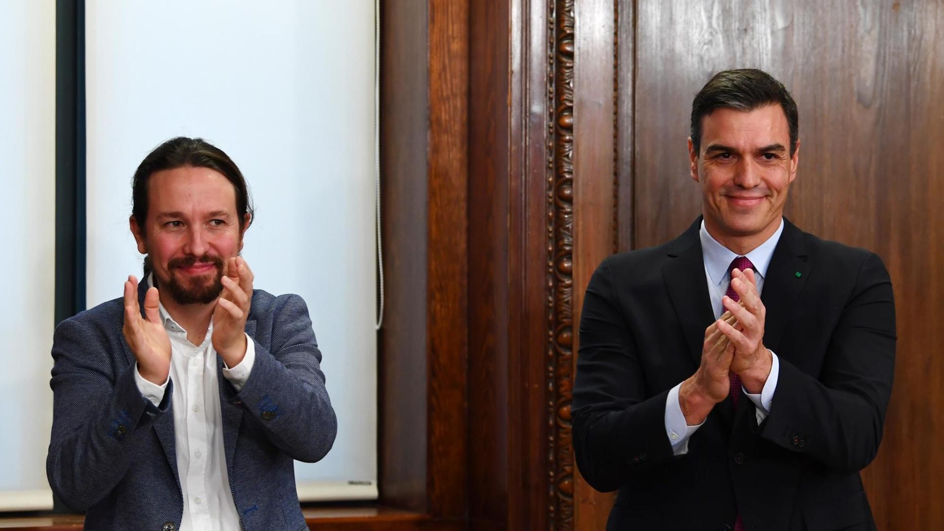 Die Politiker Sanchez und Iglesias stehen nebeneinander und klatschen in die Hände.