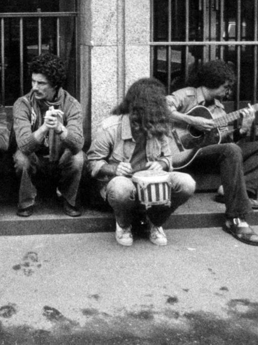 Historische Schwarzweiß-Fotografie von jungen Menschen mit langen Haaren, die auf dem Bordstein sitzen und Musik machen