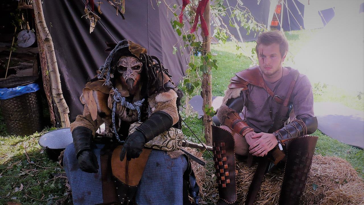 Das Bild zeigt zwei Teilnehmer des "ConQuest of Mythodea". Sie sitzen in voller Kostümierung nebeneinander vor ihren Zellten auf dem Festival-Gelände.