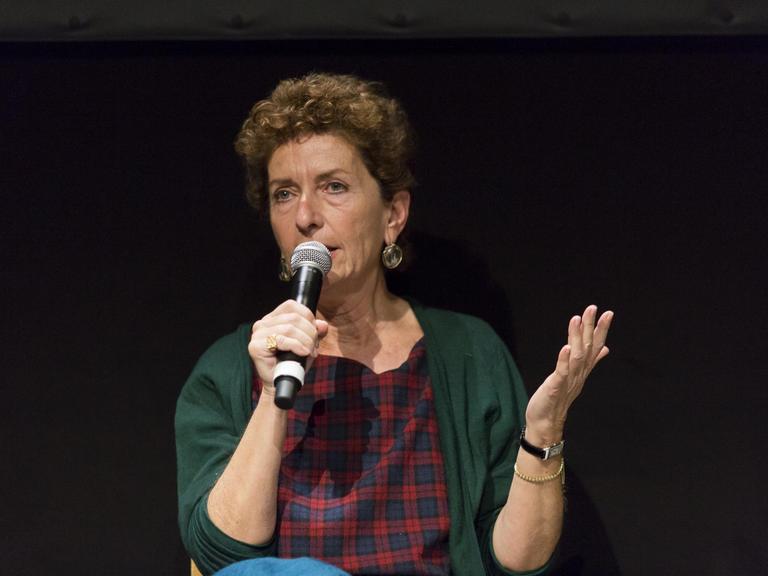Eine Frau mit dunkelblonden, kurzen lockigen Haaren. Sie trägt eine grüne Strickjacke und eine rot-blau karierte Bluse. Sie hat ein Mikrofon in der Hand und spricht. Der Hintergrund ist schwarz.