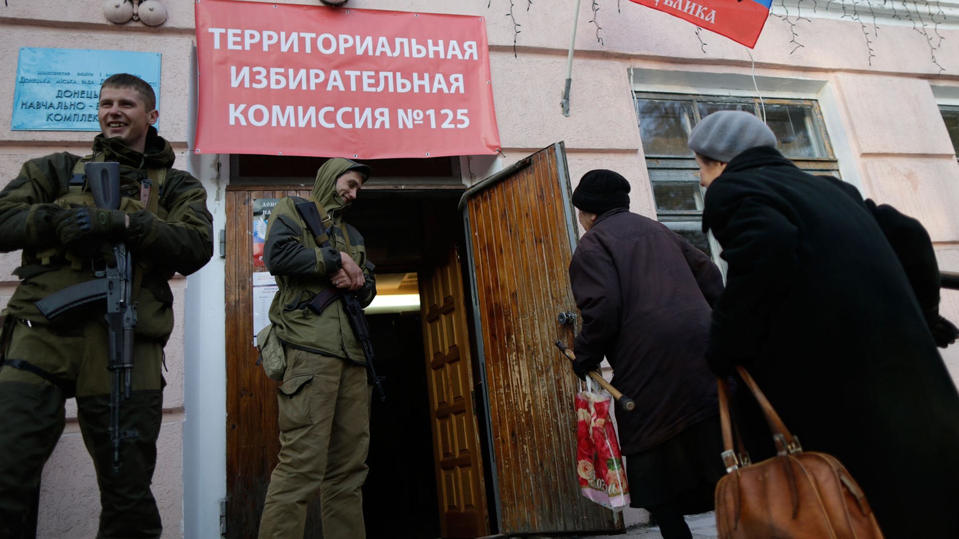 Zwei Frauen gehen in ein Wahllokal in der ostukrainischen Stadt Donezk. Zwei bewaffnete Männer in Uniformen stehen rechts und links des Eingangs
