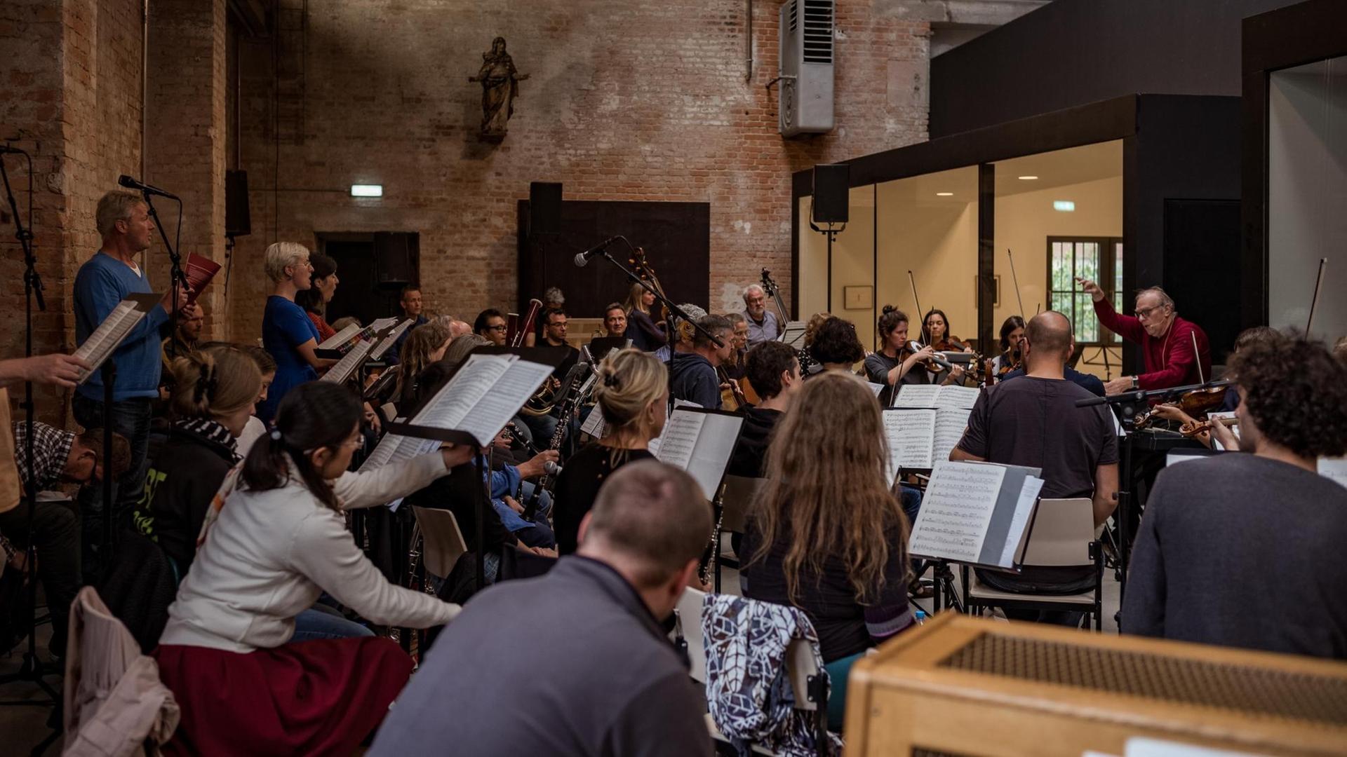 In einem Konzertsaal ist ein Orchester mit Chor seitlich zu sehen, der Dirigent steht rechts im Bild