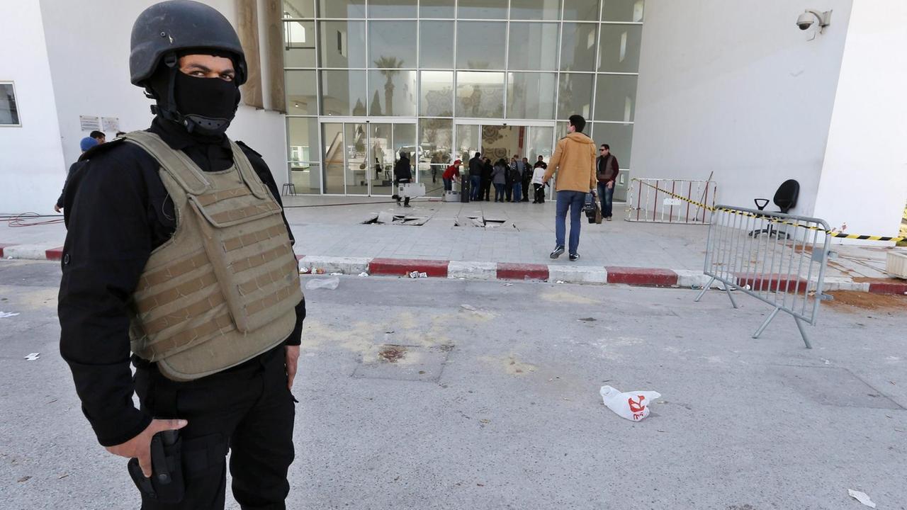Ein schwarz gekleideter Polizist mit Helm, Sturmhaube und kugelsicherer Weste steht vor dem abgesperrten Eingang des Bardo-Museums in Tunis. Hinter ihm sieht man Ermittler; rechts vom Eingang sind Blutspuren zu sehen.