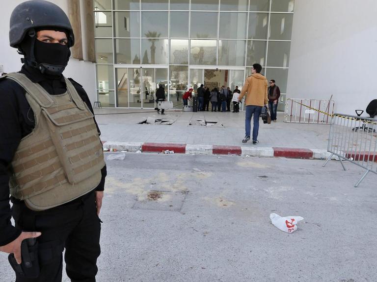 Ein schwarz gekleideter Polizist mit Helm, Sturmhaube und kugelsicherer Weste steht vor dem abgesperrten Eingang des Bardo-Museums in Tunis. Hinter ihm sieht man Ermittler; rechts vom Eingang sind Blutspuren zu sehen.