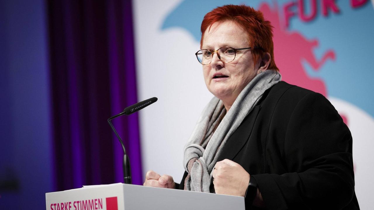 Eine Frau mit kurzen roten Haaren und einer Brille steht an einem Rednerpult und spricht in ein Mikrofon. 
