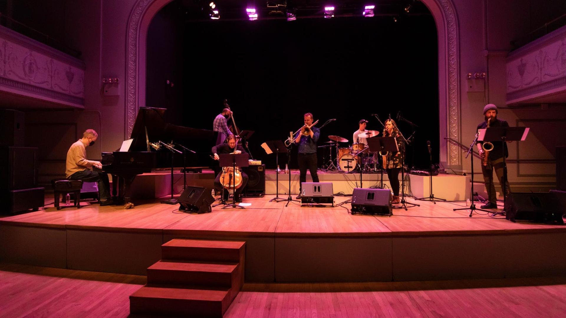 Auf einer in buntes Licht getauchten Bühne spielen eine Musikerin und sechs Musiker vor einem leeren Auditorium.