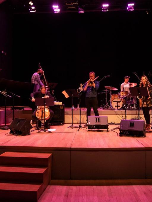 Auf einer in buntes Licht getauchten Bühne spielen eine Musikerin und sechs Musiker vor einem leeren Auditorium.