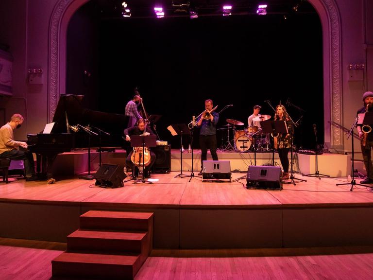 Auf einer in buntes Licht getauchten Bühne spielen eine Musikerin und sechs Musiker vor einem leeren Auditorium.