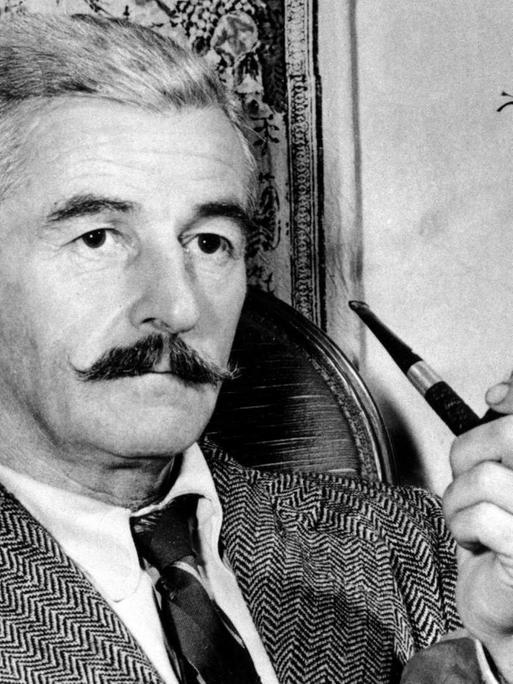 Der US-amerikanische Schriftsteller William Faulkner sitzt mit einer Pfeife in der Hand in einem Sessel