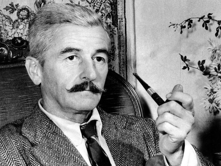 Der US-amerikanische Schriftsteller William Faulkner sitzt mit einer Pfeife in der Hand in einem Sessel