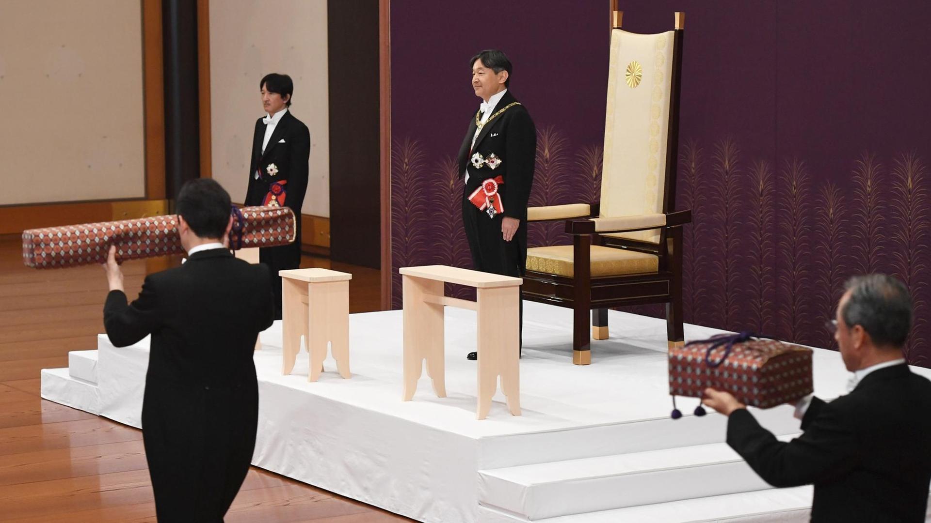 Naruhito steht vor seinem Thron. Zwei Bedienstete tragen die in zwei Paketen verpackten Insignien heran.