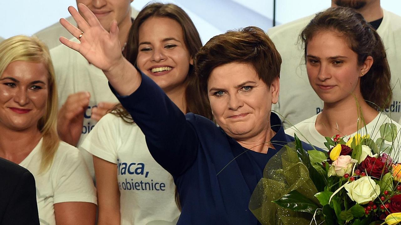 Szydlo steht mit einem Blumenstrauss vor weißgekleideten Unterstützern und winkt.