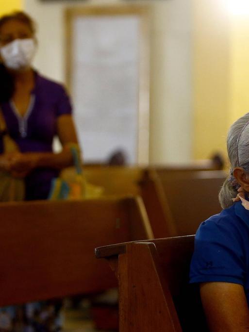 In einer Kirchenbank sitzt allein eine ältere, grauhaarige Frau. Im Hintergrund ist eine jüngere Frau mit langen dunklen Haaren und einem weißen Mundschutz zu sehen.