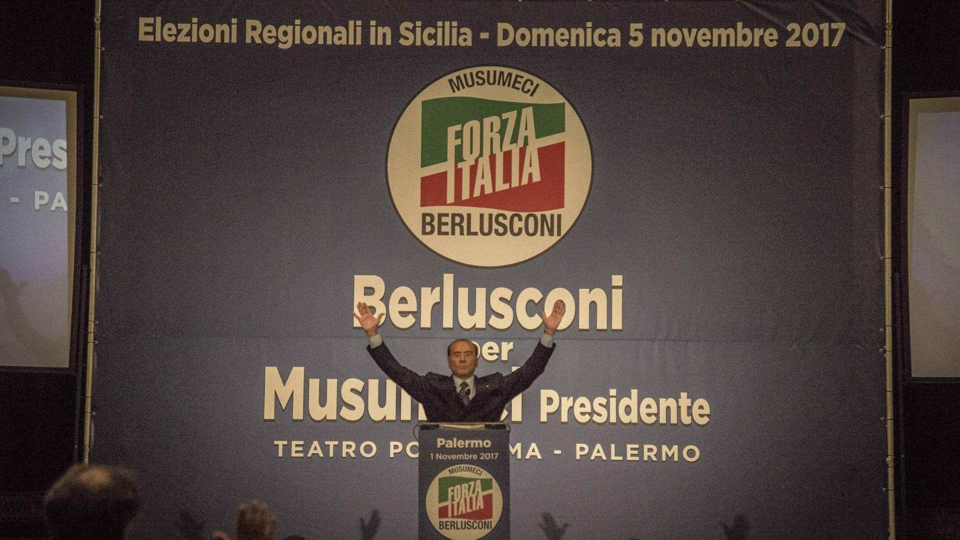 Wahlkampf in Sizilien: Silvio Berlusconi steht in Jubelpose vor seinen Anhängern. Hinter ihm stehen die Namen Berlusconi und Musumeci, der als Kandidat des Rechtsbündnisses bei der Regionalwahl auf Sizilien gewann