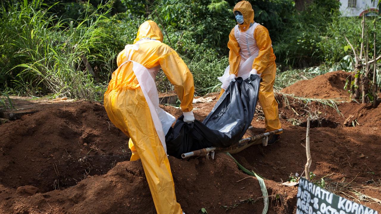 Zwei Personen in gelber Schutzkleidung heben eine in schwarze Plastikfolie verpackte Leiche in ein Grab
