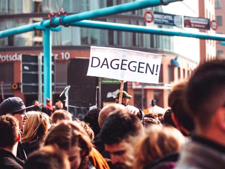 Köpfe einer Menschenmenge am Potsdamer Platz in Berlin. Es handelt sich um eine Demo. Zwischen den Köpfen ragt ein Schild mit der Aufschrift "Dagegen" hervor.