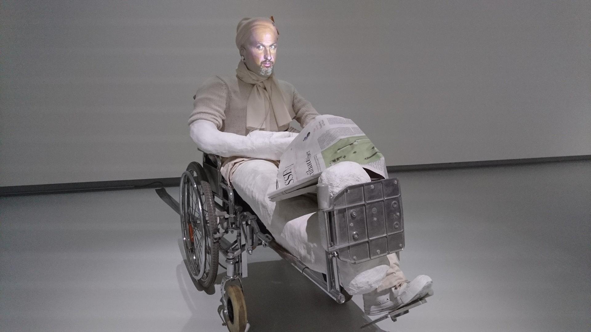 Eine in einen Rollstuhl eingegipste Puppe, auf die ein menschliches Gesicht projiziert wird.