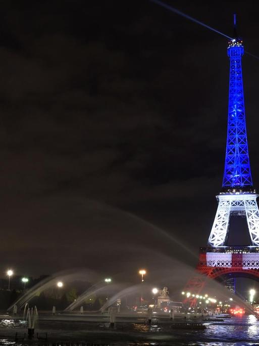 Nächtlicher Blick über die Seine auf den Eiffelturm, dessen oberer Teil blau, der mittlere Teil weiß und der untere Teil rot angestrahlt wird.