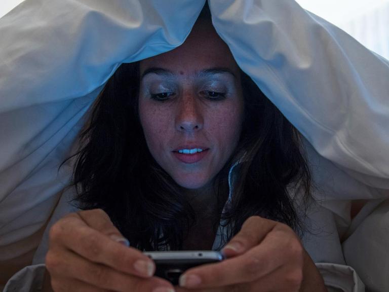 Eine Frau liegt unter der Decke im Bett und schaut auf ihr Smarphone.