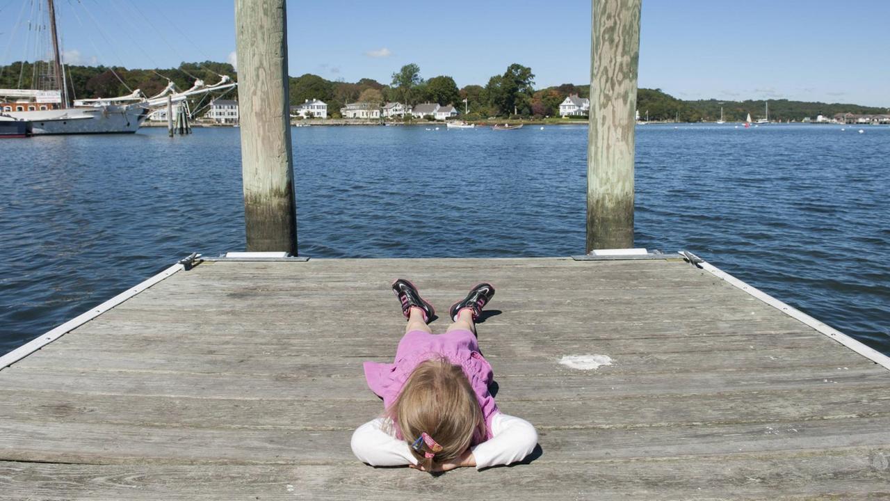 Ein Mädchen liegt entspannt an einer Mole und schaut auf ein Gewässer hinaus, auf dem Boote und Yachten sind.