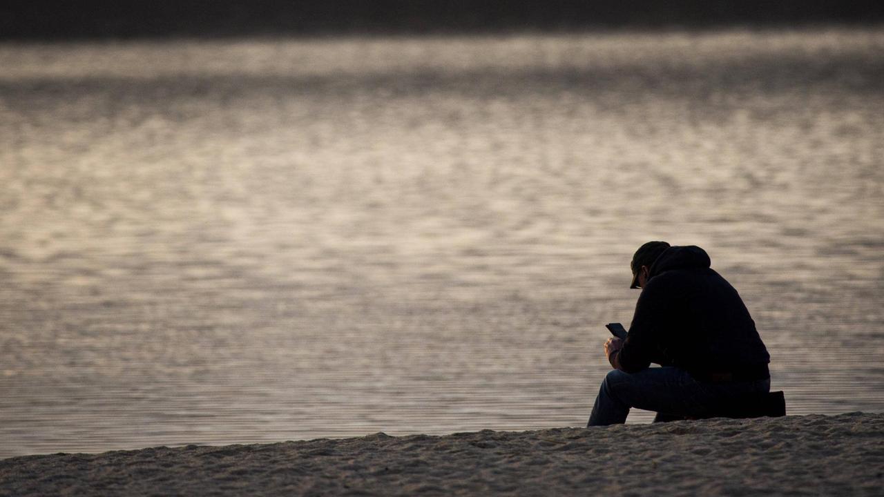 Ein Mann sitzt mit seinem Smartphone an einem ansonsten menschenleeren Strand.