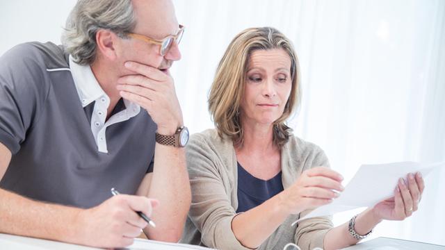 Ein Mann und eine Frau sitzen vor einem Stapel Unterlagen zusammen an einem Tisch und lesen einen Brief (gestellte Szene)