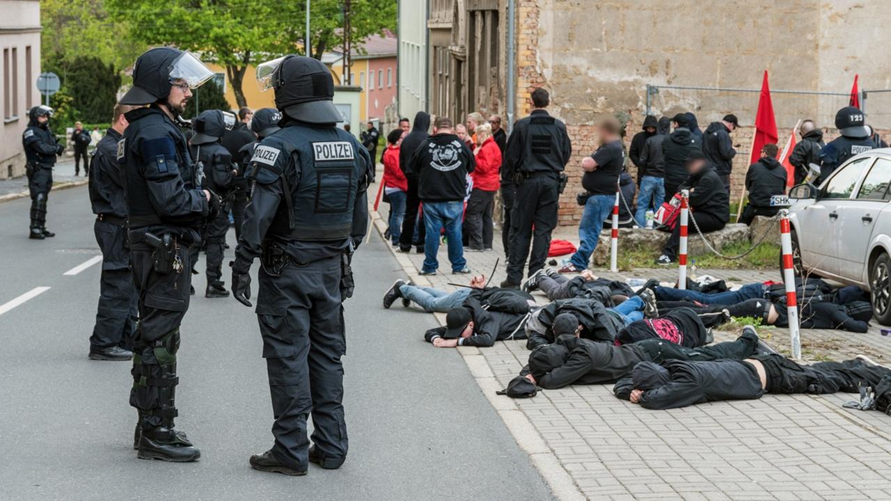 Mehrere schwarz gekleidete Männer liegen teils gefesselt auf dem Boden, andere stehen und sitzen vor einem Haus. Auf der Straße stehen viele behelmte Polizisten in schwarzer Kampfmontur.