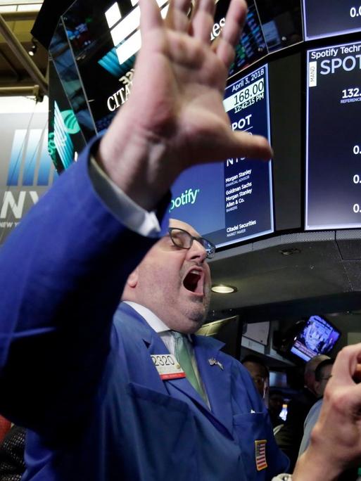Der Börsenhändler steht vor der Tafle mit dem Spotify-Kurs, zeigt alle fünf Finger der linken Hand und ruft etwas. Daneben zwei weitere Personen.