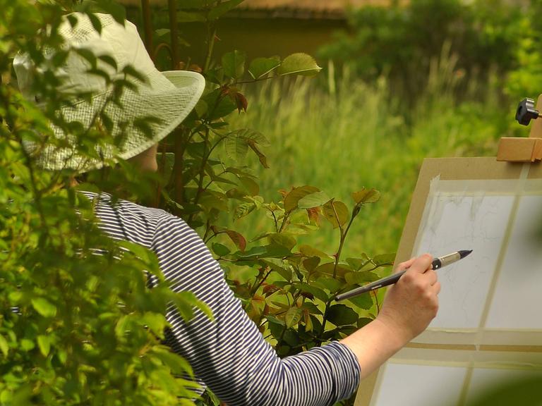Eine Künstlerin mit Hut in Rückenansicht vor einer Staffelei in einem Garten