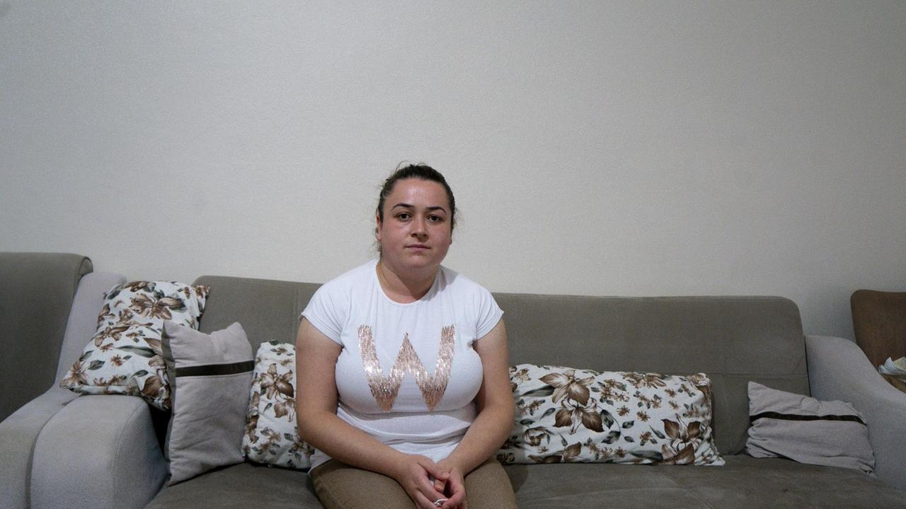 Eine junge Frau mit Pferdeschwanz und in weißem T-Shirt sitzt in einer Wohnung auf einem beigen Sofa.