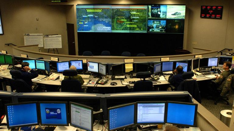 Computer und Monitore im North American Aerospace Defense Command (NORAD) auf der Peterson Air Force Base in Colorado. An der hinteren Videowand sind mehrere Landkartenausschnitte und Nachrichtenprogramme zu sehen.