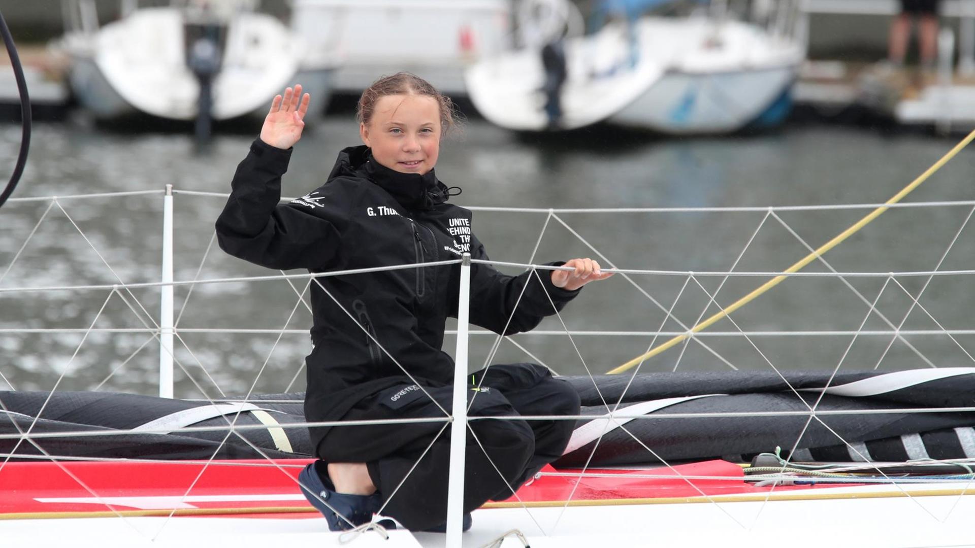 Greta Thunberg kniet an Bord, hält sich an der Reling fest und winkt lächelnd in die Kamera.