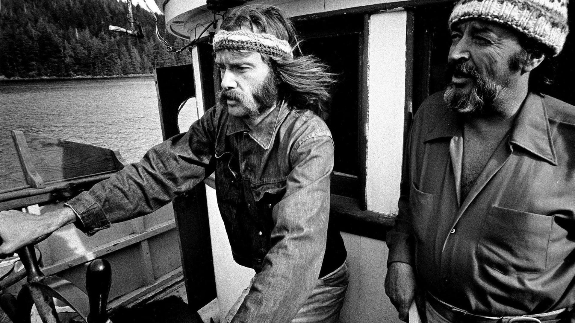 Ein Schwarzweiß-Foto von 1971 zeigt den Greenpeace-Mitgründer Bob Hunter mit langen Bart und Stirnband am Steuerrad des Kutters Phyllis Cormack