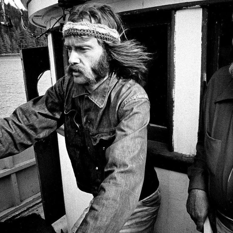 Ein Schwarzweiß-Foto von 1971 zeigt den Greenpeace-Mitgründer Bob Hunter mit langen Bart und Stirnband am Steuerrad des Kutters Phyllis Cormack 