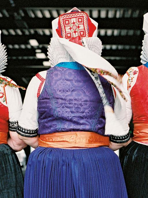 Rückansicht von drei alten Frauen in traditioneller Tracht, mit roten Hauben, weißen Spitzkragen und schwarzen oder blauen Röcken.