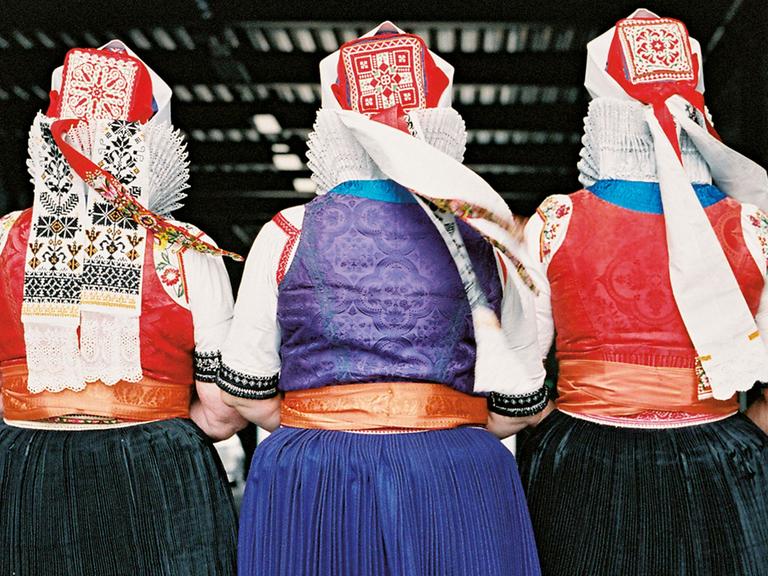 Rückansicht von drei alten Frauen in traditioneller Tracht, mit roten Hauben, weißen Spitzkragen und schwarzen oder blauen Röcken.