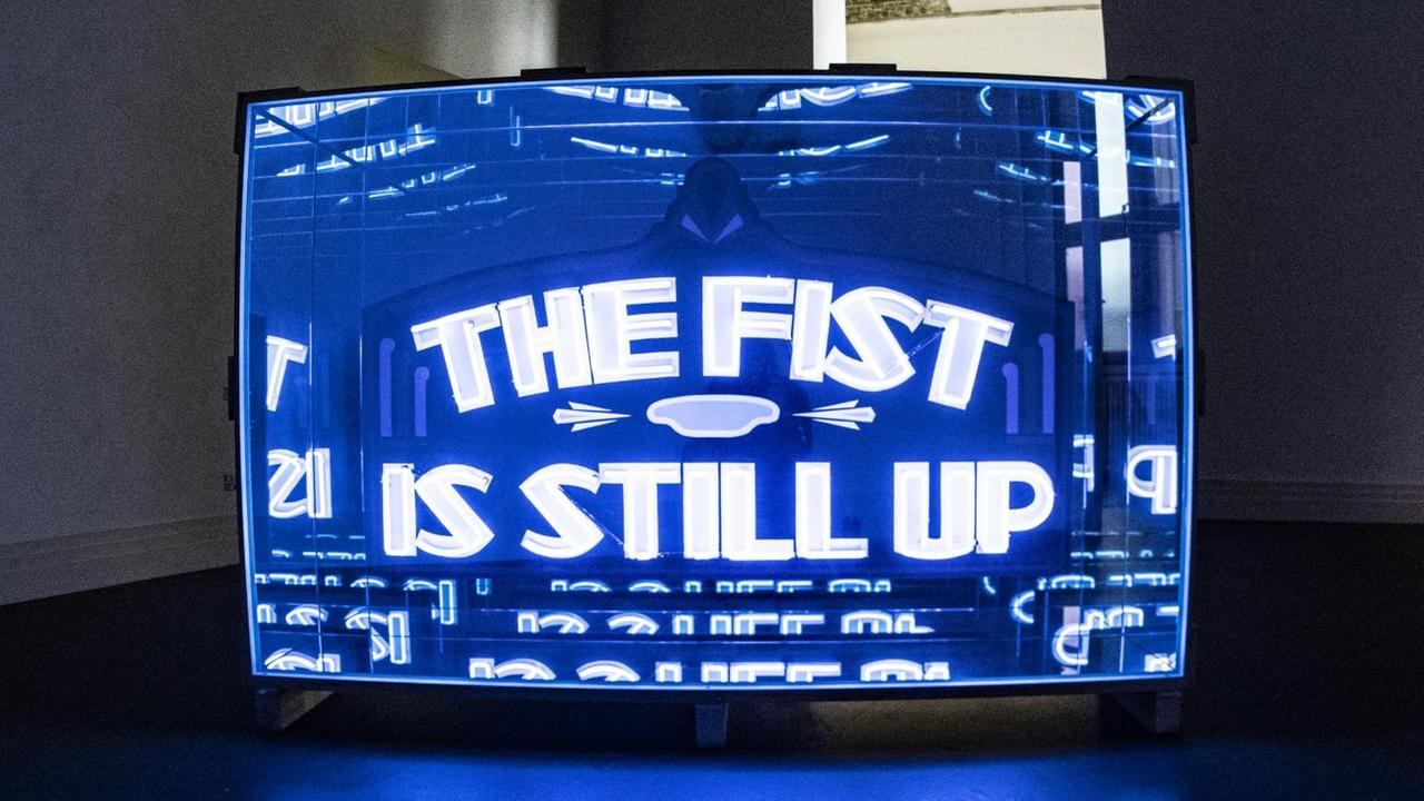 Diese Lichtinstallation, bei der auf einem blauen Leuchtschild in weißen Buchstaben "The fist ist still up" steht, ist in einem Raum im Martin Gropius Bau ausgestellt und ist Bestandteil der Ausstellung "There is no nonviolent way to look at somebody" von Wu Tsang, die vom 04. 09. bis 12.01.2020 gezeigt wird.