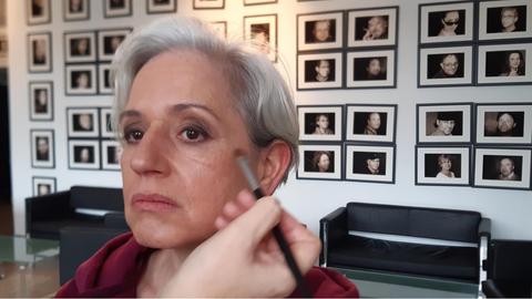 Vivian Kanner wird in der Maske für einen Fernsehauftritt. Sie tragt kurzte, graue Haare und schaut rechts an der Kamera vorbei. ´Mit einem Pinsel werden ihr Wangenknochen unterstrichen. Hinter ihr an der Wand hängen viele schwarz-weiß Portraits.