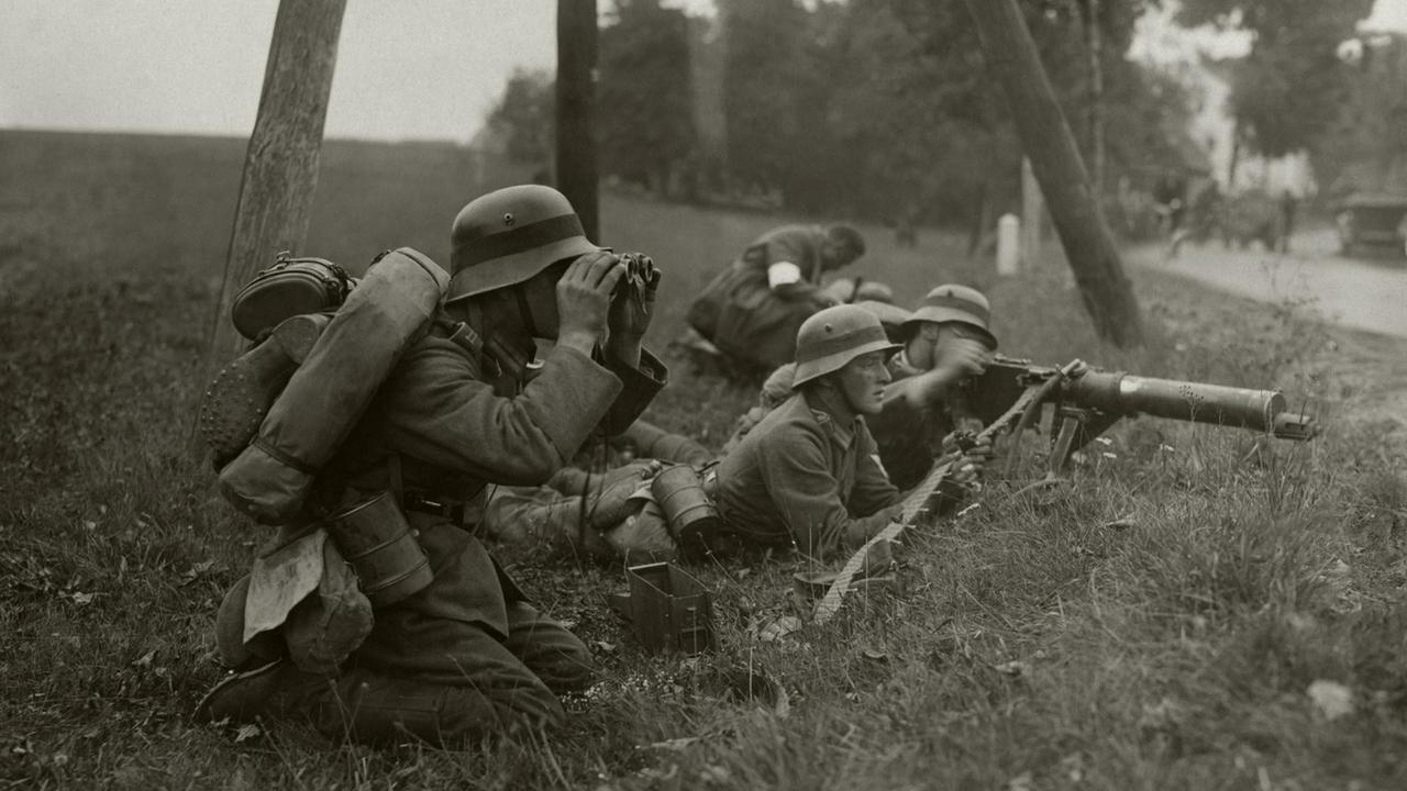 Manöver der Reichswehr an einem Straßenrand in Schlesien während der Zeit der Weimarer Republik: Ein Soldat schaut knieend in ein Fernglas, weitere liegen, einer vor einem aufgestellten Maschinengewehr